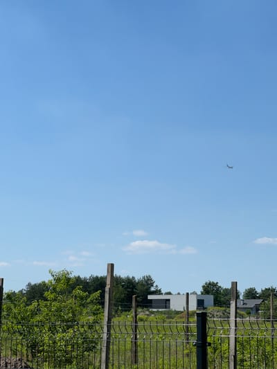 This media file depicts an outdoor scene under a clear blue sky with scattered clouds. A chain-link fence runs along the right side of the frame, with wooden posts supporting it. Beyond the fence, lush green foliage and trees are visible. In the background, a modern white building with dark accents stands amidst the greenery, suggesting a residential or commercial area. Further in the distance, more trees and what appear to be other structures can be seen.
A notable detail in the sky is a small, dark object that appears to be a bird in flight, with its wings spread. The lighting suggests it is daytime, likely morning or afternoon, given the bright sky and clear visibility. The overall impression is one of a tranquil, rural or semi-rural setting in an area that combines natural elements with modern architecture. No people are visible, and the scene does not depict any specific activity or event, but rather a static view of the landscape. No text is visible in the image. The scene appears to be located in or near Konstancin-Jeziorna, Poland, based on the provided location context.