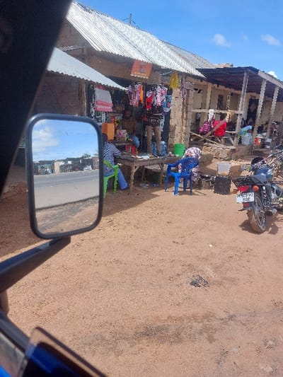An outdoor scene in Kimamba, Tanzania, depicts a commercial roadside area under a bright, partly cloudy sky. The perspective is from inside a vehicle, with its side mirror reflecting a paved road and distant landscape visible in the foreground. The immediate ground is unpaved, reddish-brown dirt with scattered darker stains.
On the right, a row of single-story commercial stalls features corrugated metal roofs and primarily exposed cinder block or wooden construction. The leftmost stall displays various merchandise, including clothing items hanging from its facade and packaged goods on shelves and a wooden table. An orange rectangular sign is affixed near the roofline of this stall.
Three adult males are present near the leftmost stall. One male in a brown shirt sits behind the wooden table, attending to goods. Another male, wearing light pants and a patterned shirt, stands upright next to the table. A third male, in a plaid shirt and blue jeans, sits on a green plastic chair at the table, facing away from the camera. A blue plastic chair is positioned nearby on the dirt ground, occupied by a partially obscured figure. Further to the right, within an adjacent open-front stall, a figure in red attire is seated.
A black motorcycle with Tanzanian license plate MC990 BQE is parked on the dirt ground on the far right. Various containers and items are scattered around the stalls, including a green bucket near the standing male. The scene suggests a local marketplace or gathering point.