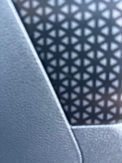 This is a close-up, abstract photograph focusing on textures and patterns. The left side of the image features a dark, perforated material with a repeating geometric pattern of interconnected triangles, creating a shadow-like effect. This pattern is out of focus, giving it a soft, almost ethereal appearance.
The right side of the image displays a different texture, appearing to be a light-colored, pebbled surface, possibly leather or a similar material. This surface is more in focus, revealing a tactile quality. A diagonal line separates the two distinct textures, suggesting a seam or an edge between different components. The overall lighting is soft, with a slight glare visible on the pebbled surface, indicating it's likely an indoor setting under artificial light or diffused sunlight. There are no people, animals, or clear signs of a specific activity. The scene is abstract and focuses on the interplay of light, shadow, and material textures. The location context of Parekklisia Community, Cyprus, does not offer specific visual cues within the frame. There is no visible text in the image.
