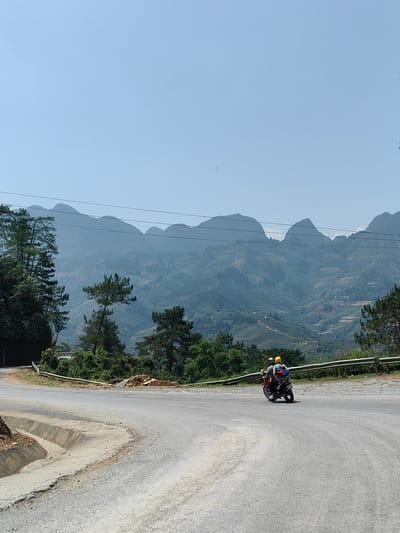 The image shows a road in Bạch Đích Commune, Vietnam, surrounded by mountains and trees. A red motorcycle with two passengers wearing helmets is turning on the road. The road has a drainage ditch on the left side. A guardrail is visible on the right side of the road. Power lines are strung across the mountain backdrop.