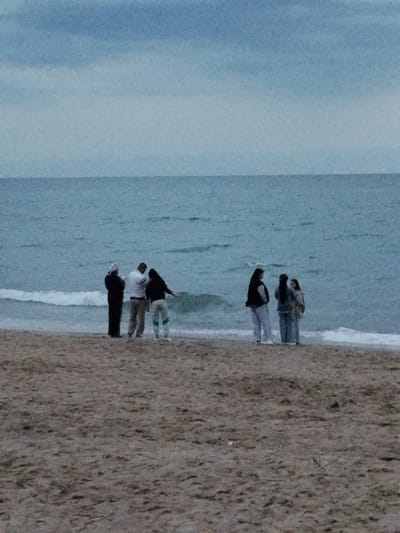 This image depicts a group of six people standing on a sandy beach next to a calm body of water under an overcast sky. The people are clustered together, appearing to be engaged in conversation or observing something at the water's edge. Three individuals are in the foreground to the left, and three are in the foreground to the right. The people on the left include a woman in a black garment, a man in a white shirt and light-colored pants, and another person in a dark top and light-colored pants. On the right, there are three women dressed in casual attire, including sweaters and pants.

The scene suggests a cool or cloudy day, possibly late afternoon or early morning, given the muted light. The waves are gentle, lapping at the shore. The background is dominated by the vast expanse of the sea and the sky, which are a uniform shade of blue-gray. The sand is a light brown, with some texture and scattered debris visible. There are no discernible objects other than the people and the natural elements of the beach and sea. The overall mood appears relaxed and contemplative. No text is visible in the image. The location is stated to be Bosteri, Kyrgyzstan.