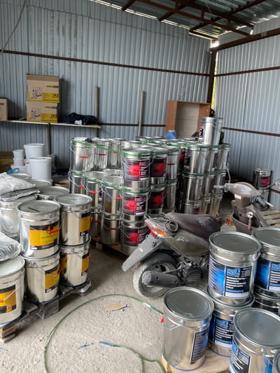 The image depicts an interior scene, likely a storage area or workshop, in Кенен Әзірбаев ауылы, Kazakhstan. The environment is industrial, with corrugated metal walls and a metal-beam ceiling. The floor is concrete. Dominating the scene are numerous large, silver metal buckets, many stacked on wooden pallets. These buckets appear to contain paint or a similar liquid product, with labels visible on some, indicating brands such as "PROTHERMA-O" and "PROFI PAINTS LTD".
In the foreground and midground, several stacks of these buckets are arranged. To the left, there are also some white plastic containers and bags of what appear to be granular material. In the background, on shelves mounted to the corrugated wall, are cardboard boxes, also with labels.
A prominent object in the midground is a scooter, parked amongst the buckets. The scooter is a brown-grey color and appears to be older. Keys are in the ignition.
The lighting suggests it is daytime, with natural light entering from unseen openings, creating some shadows. There are no people visible in the image. The overall impression is one of a commercial storage space for industrial or construction materials.