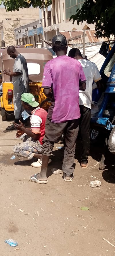 This image depicts an outdoor street scene in Gwale, Nigeria, under bright daylight. Four adult males and two three-wheeled motorized rickshaws are visible.
In the foreground, a dark-complexioned male stands with his back mostly to the camera, positioned slightly right of center. He wears a black baseball cap, a purple short-sleeved t-shirt with a textured pattern, dark trousers, and grey-and-white slide sandals. To his immediate right, partially obscured, is another dark-complexioned male, also with his back to the camera, wearing a white short-sleeved t-shirt with "Gwale" visible on the back, dark trousers, and beige flip-flops. He stands next to a blue three-wheeled rickshaw, which has "Gwale" written in white on its side.
To the left of the central figure, a dark-complexioned male crouches or squats, facing towards his clasped hands. He wears a green baseball cap, a red and white long-sleeved shirt, camouflage-patterned trousers, and white slip-on shoes. A white rectangular object is tucked into his left waistline. He wears a ring on his right ring finger.
Further left in the midground, a dark-complexioned male stands, facing slightly right, with his hands raised and clasped in front of him. He wears a light grey long-sleeved tunic over matching trousers and dark sandals. Behind him is the rear of a yellow three-wheeled motorized rickshaw.
The ground surface is unpaved, consisting of light brown dirt or soil, with scattered small debris including leaves, small sticks, a blue plastic wrapper, and small white rocks. Shadows are distinct, indicating clear weather. The background features light-colored multi-story buildings with various architectural details and some corrugated metal roofing under a clear sky. Dark green foliage from trees is visible in the upper sections of the image, providing overhead cover.