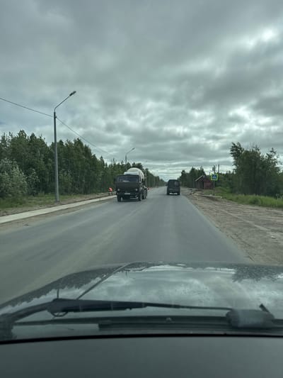 This media file depicts a view from inside a vehicle, looking out at a road with overcast skies. The scene appears to be along a rural or semi-rural road, with trees lining the left side. In the distance, there are at least two trucks visible on the road. The foreground shows a portion of the vehicle's interior, including what appears to be a windshield with a wiper blade. The road surface is wet, suggesting recent rain or damp conditions.

The subjects in the image are primarily vehicles and the natural environment. There are multiple trucks, one of which is a cement mixer truck, indicating construction or transport activity. A few streetlights are visible along the side of the road. The sky is heavily clouded, with patches of brighter light suggesting diffused sunlight breaking through. The time of day is unclear due to the overcast conditions, but it does not appear to be early morning or late evening.

Notable details include the wet road, the presence of construction equipment (cement mixer truck), and the overcast, potentially damp weather. The general mood is somewhat subdued due to the weather, but the presence of traffic suggests ongoing activity.

There is a yellow pedestrian crossing sign visible on the left side of the road, with a blue square sign below it. The license plate on the cement mixer truck is partially visible, and it appears to have Cyrillic characters, consistent with a Russian location.

The location context provided is Parma, Russia. This aligns with the presence of Cyrillic script and the general landscape. The activity appears to be everyday travel or transport on a road that might be undergoing some form of maintenance or construction, given the presence of the cement mixer.