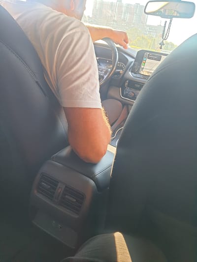 The image shows the interior of a car from the perspective of a passenger in the back seat. The focus is on the driver, a person with a beard wearing a white t-shirt, whose right arm rests on the center console. The driver's hand is visible on the steering wheel. The car's dashboard features a touch screen displaying navigation, a climate control panel, and a digital speedometer. A beaded rosary with a cross hangs from the rearview mirror. The back seat area shows air vents and USB ports. The scene appears to be set in an urban environment, with buildings and trees visible through the windshield. The lighting suggests it is daytime, possibly late afternoon due to the warm sunlight casting shadows.