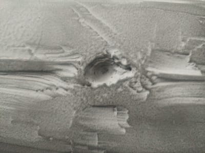 The image shows a close-up, grayscale view of a rough, sandy surface with a circular indentation. The surface appears textured with visible striations and granular material. The indentation is roughly oval-shaped and appears to be a depression or cavity in the sand. The lighting creates shadows within the indentation, suggesting depth. The overall scene is devoid of any human presence or discernible city elements, making it impossible to ascertain a location within Тлянче-Тамак, Russia. The image is purely an abstract representation of a textured surface with an indent.