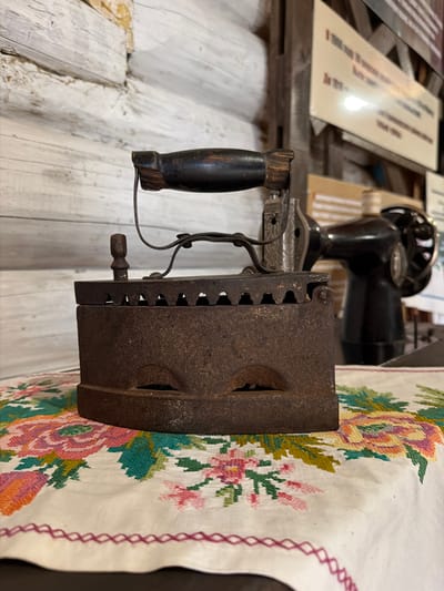 An antique, cast-iron flat iron, heavily oxidized, rests on a white cloth embroidered with colorful floral patterns. The iron has a dark wooden handle and a metal support mechanism. Behind the iron, a vintage black sewing machine is visible, also out of focus. The background consists of weathered wooden walls and a light-colored sign with text. The scene appears to be set in a room, possibly a museum or historical display, in Ust'-Kabyrza, Russia.
