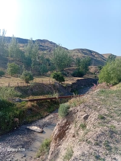 A wide-angle, low-angle outdoor shot depicts a dry, rocky stream bed with a shallow, murky stream flowing through it. The stream bed is lined with dark grey rocks and gravel, with some sparse green vegetation growing on its banks. A large, light-colored rock is partially submerged in the stream. A rusty metal pipe runs horizontally across the stream bed. The background features rolling hills covered with dry grass and scattered trees. The sky is clear and blue. In the bottom left corner, a watermark reads "Galaxy A06". The scene appears to be located in Kojo-Aryk, Kyrgyzstan.