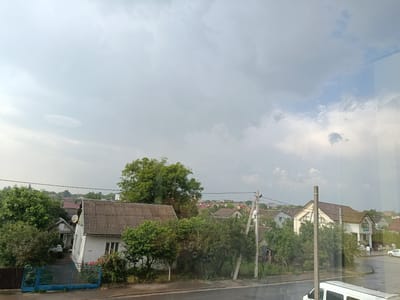 The image depicts a residential street in Pustomyty, Ukraine, under an overcast sky with scattered clouds. The view is from an elevated perspective, looking down onto a road that curves to the right. On the left side of the road, several houses are visible, characterized by their pitched roofs, some with grey corrugated material and others with brown tiles. A blue metal gate stands in the foreground on the left, leading to a yard with a white house and a small car. Lush green trees and bushes are abundant, surrounding the houses and extending into the background, creating a somewhat overgrown appearance. Power lines are strung across the scene, supported by concrete poles.
Further down the street, on the right side, more houses are visible, including a larger white building with a prominent brown-tiled roof, and another building that appears to be a shop or commercial establishment with a sign featuring blue text. A white vehicle, possibly a van or minibus, is parked in the foreground on the right, partially obscuring the view of the street. The road surface appears wet, suggesting recent rain. The overall impression is of a quiet, suburban neighborhood on a cloudy day.
