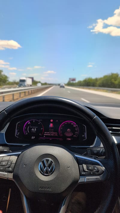 The image is a first-person perspective from the driver's seat of a car on a highway. The steering wheel, a black leather Volkswagen model with the brand's logo in the center and "AIRBAG" written below, is prominently in the foreground. The digital dashboard displays various information, including a speed of 134 km/h, a fuel efficiency reading of 6.5 l/100km, and song title "HIP HOP, AFROBEATS, AMAPIANO, R&B LIVE...". The road ahead is visible, with other vehicles in the distance and trees lining the sides. The sky is a clear blue with scattered white clouds. The context of Shtarkovo, Bulgaria is not explicitly depicted or derivable from the visual information.