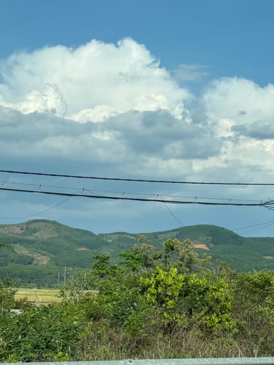 The image depicts a landscape near Đăk La, Vietnam. The foreground contains power lines running vertically, partially obscuring the view. To the left, a sky with white cumulus clouds against a blue background is visible. The right side of the image shows a series of green mountains, with areas of exposed reddish soil visible on some slopes. In the foreground, foliage and trees are interspersed with a possible rice field area.