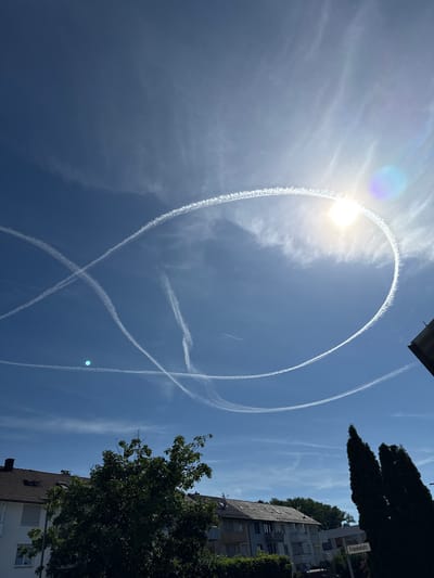 In Salach, Germany, on a clear, sunny day, the sky is filled with numerous contrails from airplanes. A prominent circular contrail is visible in the upper right quadrant of the image, partially obscured by the sun which creates a lens flare. Several intersecting straight contrails also cross the sky. The lower portion of the image shows residential buildings with tiled roofs and trees. A street sign reading "Fränkelstr." is visible on the right side, indicating the location is likely on or near Fränkel Street in Salach. The scene is static, with no people or animals directly visible.