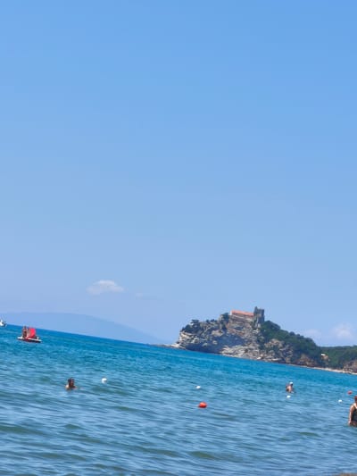 The image depicts a sunny day at a beach in Castiglione della Pescaia, Italy. The clear blue sky and calm turquoise water suggest pleasant weather, likely mid-day. In the foreground, several people are enjoying the sea, with some swimming and others wading near the shore. White buoys and a few orange ones are scattered across the water, marking an area within the sea.
Further out, a small red sailboat with two people on board is visible. In the middle ground, a rocky island rises from the sea, crowned by an old stone building, possibly a fortress or castle, with a reddish-tiled roof. The island is covered in lush green vegetation. The distant shoreline shows hills and more greenery under the bright sky.
The overall scene conveys a sense of leisure and relaxation, typical of a summer vacation. There is no visible text in the image. The lighting indicates it is daytime, with the sun casting bright light and creating reflections on the water.