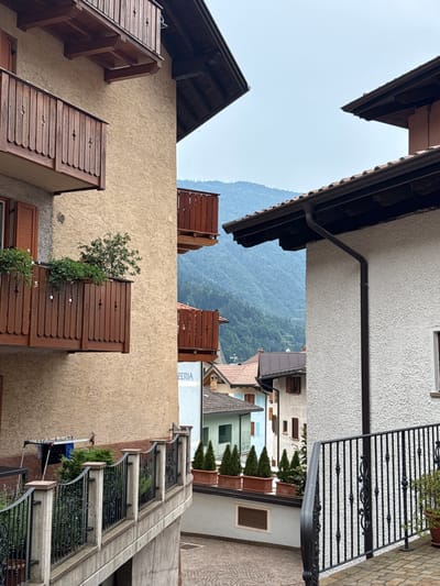The image shows a view down a narrow street in Molveno, Italy, with buildings on either side and a mountainous landscape in the background. The street is paved with cobblestones. On the left, a building with a textured beige facade features multiple wooden balconies with ornate railings and planters containing greenery. A clothesline with laundry is visible on one of the lower balconies. On the right, a white stucco building with a dark tiled roof and a dark downspout is prominent. A black metal railing lines a set of stairs leading up to this building. In the distance, several other buildings with tiled roofs are visible, and beyond them, a lush green mountain range under a hazy sky. The overall scene depicts a charming, traditional Italian village setting.