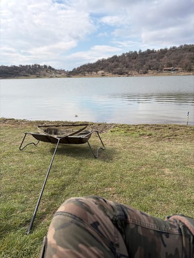 The image displays a first-person perspective of a lakeside scene in Cherna Gora, Bulgaria. In the immediate foreground, the knees and thighs of a person wearing camouflage-patterned trousers are visible. Directly ahead, on a patch of sparse green grass, rests a dark green fabric fishing cradle or unhooking mat, supported by a metallic four-legged frame, with an additional stabilizing pole extending towards the camera. A mesh net is visible within the cradle. To the right, near the water's edge, a green and yellow marker post with the numeral "5" is partially submerged. The mid-ground is dominated by a calm body of water, reflecting the sky and distant features. The far bank is lined with a density of bare deciduous trees or shrubs, suggesting a cooler season. Scattered along the distant bank are a small white structure, a larger building with a blue roof, and a wooden gazebo. The sky above is partially cloudy with patches of blue visible through white and grey clouds.