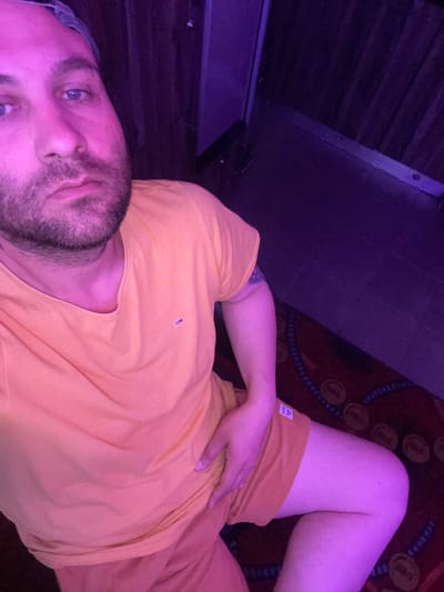 The image shows a man sitting down with his left hand on his thigh. He is wearing an orange t-shirt and orange shorts. The man has blue eyes and a beard. He is looking at the camera. The background is dark with purple lighting. There is a carpet on the floor with a circular pattern. The image does not contain any identifiable information about the city of Newry, United Kingdom.