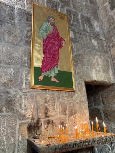 An icon depicting a reclining figure in a red robe hangs on a gray stone wall. The painting has a gold frame. A niche in the wall is visible above the icon. To the right, a stone structure with lit candles protrudes from the wall, the flames emitting a warm glow. The wall is constructed of roughly hewn stones of varying sizes.
