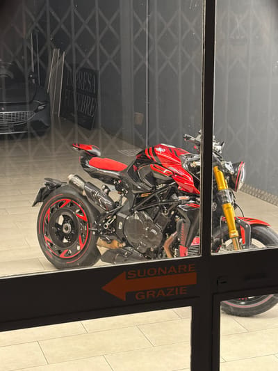 An exterior shot shows a red and black motorcycle parked inside a building with a dark, corrugated metal grate in the background. A black car is partially visible to the left of the motorcycle. A sign with "IMPRESA FUNEBRE" in large, capital letters is visible on the wall behind the car. A dark horizontal panel with orange text that reads "SUONARE GRAZIE" with an orange arrow pointing left is in the foreground, obscuring the lower part of the motorcycle. The floor appears to be tiled. The image suggests the motorcycle is parked in an indoor showroom or garage, possibly associated with a funeral home in Novara, Italy, given the text on the sign.