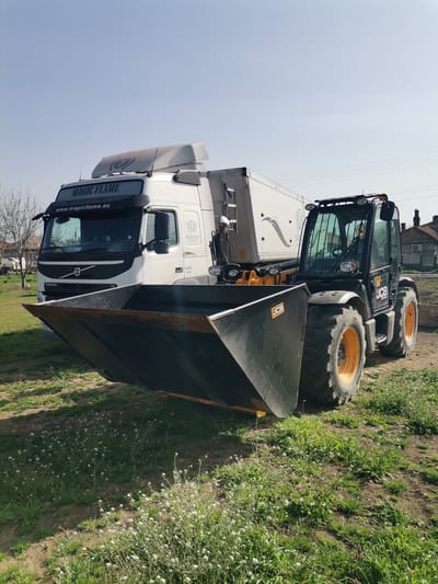 The image shows a white Volvo semi-truck with "MAGIC FLAME" printed on the visor and "www.magicflame.eu" underneath, coupled with a large silver or white trailer. In front of the truck is a black JCB telescopic handler with a large, dark-colored bucket attachment, sitting on a patch of grass and weeds. The JCB has black and yellow wheels. The background includes a pale blue sky, a tree without leaves, and a residential building with a red tile roof. Based on the location, this scene is likely in or near Paisievo, Bulgaria.