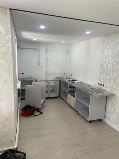 The image shows an unfinished kitchen under renovation. The room is a corner of a residential apartment, likely in the city of Alagir, Russia, given the context. The walls are partially covered with a textured wallpaper and appear to have some unfinished plastering. The ceiling is white with recessed lighting fixtures.
The primary subjects are kitchen cabinets in various stages of assembly. On the left, there are several grey base cabinets, some open and some with doors. In the foreground, to the left of the cabinets, is a red tool bag, open and containing tools.
Along the back wall, a long countertop, covered in protective plastic, has been installed over a row of white and grey cabinets. This countertop has cutouts that suggest future placements for a sink or stovetop. Several electrical outlets are visible on the wall behind the countertop.
The floor is covered with light-colored laminate flooring. Some pipes are visible along the left wall, indicating plumbing. There are no people present in the image. The overall impression is a room in the process of being outfitted as a kitchen.