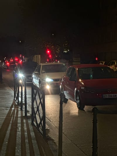 The image depicts a nighttime street scene in Colombes, France, showing stationary vehicles on an asphalt road. In the immediate foreground on the left, a black metal fence with decorative criss-cross segments and vertical posts borders a concrete sidewalk, casting elongated shadows to the right. A red Volkswagen hatchback, bearing license plate EY-394-GM, is stopped on the right side of the road, oriented towards the left. Behind it, a silver sedan is stopped, with its headlights brightly illuminated. Further back, a dark-colored vehicle also displays illuminated headlights. In the background, multiple red traffic lights are visible, indicating the vehicles are stopped at an intersection or in traffic. Additional vehicles are discernible in the distance by their illuminated red taillights. The overall environment is dark, with faint outlines of trees and buildings in the background. No people are present.