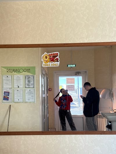 The image is a reflection in a mirror. The mirror is hanging on a wall with textured beige wallpaper. The wall also has a poster with the Cyrillic text "РПИАМДОФИН" on a green background and a collection of certificates. Above the poster, there is a sticker commemorating the years 1941-1945.
In the reflection, two individuals are visible. One is wearing a dark jacket and grey pants, looking at a phone. The other is wearing a red jacket and dark pants, also holding a phone. Behind them is an exit sign labeled "ВЫХОД" in Cyrillic, and a door. Beyond the door, there is a view of the outside, possibly a cityscape. The reflection also includes a white sink, a towel dispenser and ceiling lights. The overall scene appears to be an indoor space, possibly a hallway of a building. Since it is not possible to identify the location as the city of Gory, Russia from this image, there is no way to tell whether it is there.