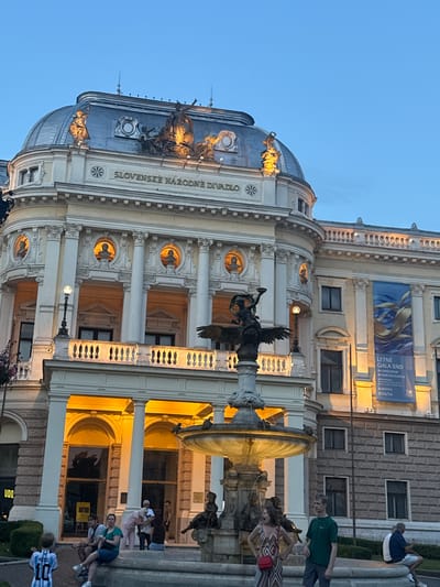 This image captures the Slovak National Theatre in Bratislava, Slovakia, at dusk. The neoclassical facade of the theatre is illuminated, highlighting its dome, columns, and balconies. A fountain with a statue of a winged figure stands in the foreground. Several people are visible in the scene, some sitting near the fountain and others standing. The sky is a deep blue, indicating the evening hour. A banner on the side of the building displays text related to a summer gala.