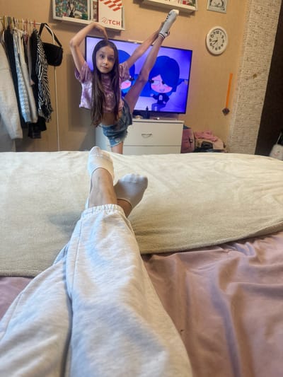 The media file depicts a young girl in a room, seemingly in a casual or playful moment. She is lying on her back on what appears to be a bed or sofa, with her legs extended upwards. She is wearing a light purple tie-dye t-shirt and denim shorts, with white socks on her feet. Her hair is long and dark brown, and she is looking directly at the camera with a neutral expression.
In the background, a television screen is visible, displaying an animated character with blue hair. To the left of the television, a round wall clock shows the time to be approximately 3:50. There are also some framed pictures on the wall, a white cabinet, and a stack of clothes on a rack. The overall setting suggests a domestic environment, possibly a bedroom or living room.
The lighting appears to be indoor artificial light, and there are no clear indicators of the time of day or weather conditions outside. The mood seems relaxed and informal, with the girl posing for the camera. The image is oriented vertically.
Visible text includes "TCH" on a red and white box to the left of the television and numbers on the clock face.
