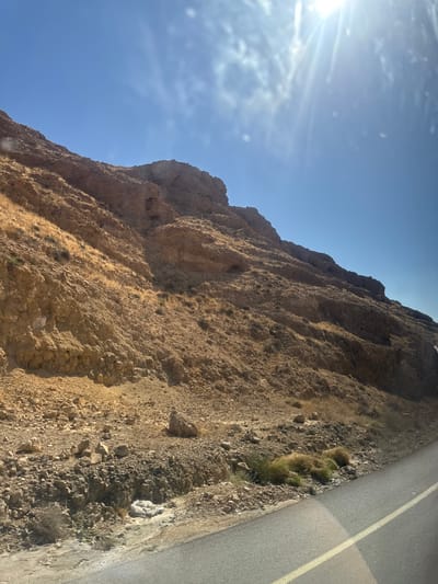 A low-angle shot shows a craggy, brown, sunlit hillside in Arad, Israel. The sun shines brightly in the upper left of the frame, against a clear blue sky. The hillside appears rocky and uneven, with varying textures and shadows. At the right edge, a paved road with a yellow line runs parallel to the hillside. Patches of dry, brown grass grow along the edge where the hillside meets the road. No people or other objects are visible.