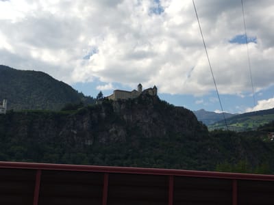 The image shows a view of the Säben Monastery, also known as Sabiona Abbey, located on a rocky outcrop above the town of Klausen (Chiusa), in South Tyrol, Italy. The monastery complex, with its distinct red-tiled roofs and towers, is perched prominently on the hillside, surrounded by lush green trees and steep, rugged terrain. The sky above is partly cloudy, with a mix of white clouds and blue patches. In the foreground, a portion of a dark, reddish-brown structure, possibly a fence or barrier, is visible, running horizontally across the bottom of the frame. The overall scene depicts a historical and picturesque landscape, characteristic of the Alpine region.