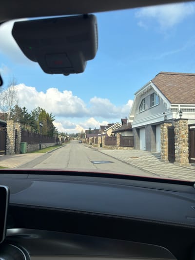 The image shows a residential street in Selyatino, Russia, viewed from inside a car. The street is lined with houses featuring stone facades and brown roofs. A sidewalk runs along the right side of the street. The sky is blue with white clouds. A car dashboard and a rearview mirror are visible in the foreground.