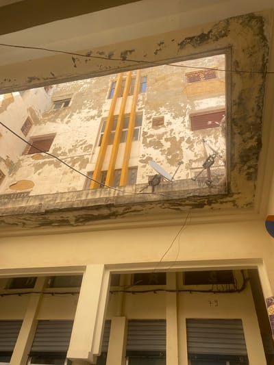 The image shows an exterior view of two adjacent buildings in El Hajeb, Morocco. The foreground building is painted a light yellow and features multiple storefronts with closed, corrugated metal shutters. Above the storefronts are rectangular windows. Several electrical conduits and wires are visible running along the exterior wall. The number "79" is spray-painted on the wall between two of the windows.
Behind the foreground building, a second, older building is visible. This building has a weathered facade with peeling paint, revealing patches of older plaster and brickwork. It features numerous windows, some with red shutters, and a decorative wrought-iron balcony. A prominent vertical element, painted a bright yellow, is present on the facade of this older building, with several narrow windows set into it. Two satellite dishes are mounted on a ledge of this older building.
The overall impression is of an urban environment with a mix of older and newer architecture, showing signs of age and wear. The lighting suggests it is daytime, with strong shadows cast by the sun. No people are visible in the image.