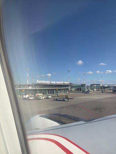 The image depicts an airport scene captured from an aircraft window on the tarmac of EuroAirport Basel Mulhouse Freiburg, located in Saint-Louis, France.

In the immediate foreground, the white wing of the originating aircraft is visible, featuring prominent red stripes, along with its metallic-grey engine nacelle. The midground presents the asphalt tarmac, marked with red painted lines. Multiple pieces of ground support equipment are parked, including several white baggage tractors, a mobile passenger stair unit, and a cargo loader. A single individual wearing a yellow high-visibility vest is faintly discernible near the terminal building, positioned beneath a gate designation, partially visible as "2BA".

The background is dominated by the multi-story terminal building, constructed of glass and concrete, displaying the illuminated white text "EuroAirport BASEL MULHOUSE FREIBURG" across its facade. Several tall, red-and-white striped poles, consistent with airfield lighting or navigational aids, stand around the terminal structure. Further to the right along the terminal, a jet bridge extends towards an aircraft, and the vertical stabilizer of another aircraft is partially visible. The environment is characterized by a clear blue sky with scattered small, white cumulus clouds, and a faint contrail is present in the upper right quadrant. No overt actions or interactions between individuals are clearly depicted beyond the presence of ground personnel and equipment.