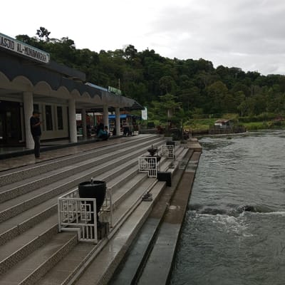 This image depicts the exterior of a mosque named "MASJID AL-MUNAWARRAH" situated beside a flowing river. The mosque's architecture features a long, covered walkway with arched supports and a prominent sign displaying its name in blue lettering. In front of the mosque, a wide, tiled staircase leads down towards the riverbank.
Several people are visible in the scene. A man in a dark shirt and green cap stands on the tiled area in front of the mosque, smoking and looking at his phone. Further down the stairs, a few individuals are seated. On the riverbank, near a small rustic wooden structure and a grassy area, a couple of people are walking. The river itself is wide and appears to have a moderate current, with some small rapids and rocks visible. The background consists of a lush, green hillside covered in dense trees.
The weather appears overcast, with a cloudy sky. The overall mood is tranquil and somewhat subdued, possibly due to the weather. The location is identified as Blang Bebangka, Indonesia. Notable objects include decorative white railings along the stairs, large dark urns, and a small wooden shack on the river.