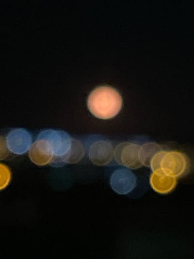 The image is a blurry, out-of-focus photograph taken at night. On the left side of the image, a single, large, soft orange-colored circular shape is visible. To the right, a vertical line of smaller, overlapping blurry circles extends from the top to the bottom of the frame. These circles alternate in color, appearing yellow, blue, and grey. The background is entirely dark, obscuring any further details about the environment. There is no discernible information in the image to definitively locate it in Rojales, Spain or to identify any people, subjects, objects, actions, or interactions.