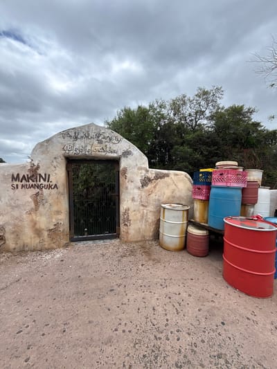The scene depicts an outdoor area in Bay Lake, United States, featuring a weathered stone archway with an iron gate and various storage containers. The archway, light in color with visible erosion and dark markings, contains a closed, dark metal gate with vertical bars. Above the gate, black Arabic script is painted, and to the left, the words "MAKINI. SI KUANGUKA" are painted in black. To the right of the archway, an assortment of plastic and metal barrels, along with plastic crates, are stacked. Visible barrels include red, blue, white, and yellow plastic types, alongside at least one white metal barrel with rust, and a prominent red metal barrel in the foreground. Plastic crates, primarily pink, blue, and yellow, are stacked on top of some barrels. The ground is a rough, light brown or reddish-brown paved surface with embedded small stones. In the background, dense green trees and foliage are visible under an overcast, gray sky. No individuals are present.