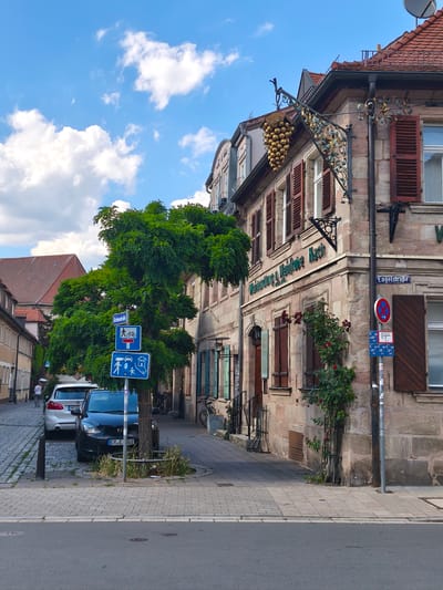 The image captures a street scene in Erlangen, Germany, with a focus on a historic building and its surroundings. The building, constructed from stone, features wooden shutters and a decorative sign depicting a bunch of golden grapes, indicating a winery or establishment related to wine. Above the entrance of the building, text reads "Weinhandlung u. Mälzerei Nach". A street sign visible on the right side of the building indicates "Engelstraße".
In the foreground, a narrow street paved with cobblestones runs alongside the building, with a sidewalk also paved in a similar fashion. A large, leafy green tree is positioned on the sidewalk, partially obscuring some traffic signs. One sign displays pedestrian and bicycle symbols with a "dead end" symbol, while another shows symbols for cars, houses, and people, suggesting a residential area or family-friendly zone.
Two cars are parked on the street. The car closer to the viewer is a dark-colored sedan with the license plate "ER E 002". A white SUV is parked behind it. On the sidewalk, a bicycle is leaning against the building.
The environment appears to be a sunny day with a bright blue sky dotted with fluffy white clouds. The architecture and street design suggest an older, well-preserved European city. The overall impression is one of a charming, traditional urban setting.