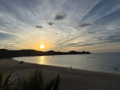 This is a wide-angle, eye-level, static shot of a beach scene at sunset in Song Cau, Vietnam. The sun is low on the horizon, casting a warm glow across the sky and reflecting on the calm ocean. The sky is filled with streaks of clouds, with some darker, more defined clouds present. In the foreground, there is a sandy beach with some palm fronds visible in the lower corners. Further back, there are several people walking on the beach and swimming in the shallow water. Small boats are scattered across the water, and a red car with a roof rack is parked on the sand near the water's edge. The background features a silhouette of rolling hills, characteristic of the Song Cau coastline. The scene evokes a peaceful and serene atmosphere.