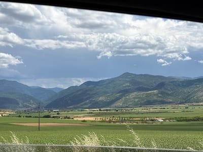 The image presents a wide-angle view of a rural landscape, likely in or near Isperihovo, Bulgaria, captured from a moving vehicle. In the foreground, a vibrant green field of crops, possibly sunflowers or a similar low-lying plant, occupies the lower portion of the frame, with wispy dry grass blades visible along the bottom edge, suggesting a barrier or edge.

Midground features include several fields with varying shades of green and golden-brown, indicating different crops or stages of growth. A single, weathered wooden utility pole stands prominently in the middle of one of these fields. Further back, a line of trees creates a natural boundary, behind which lies a cluster of buildings that appears to be a small village or settlement nestled in the valley.

The background is dominated by rolling, verdant mountains, their slopes covered in dense vegetation. The sky above is dramatic, with a mix of bright white cumulus clouds and darker, heavy storm clouds, hinting at impending or recently passed rain. The lighting is diffused due to the cloud cover, casting subtle shadows across the landscape. The overall impression is one of a vast, agricultural expanse under a dynamic sky, with the mountains providing a scenic backdrop.