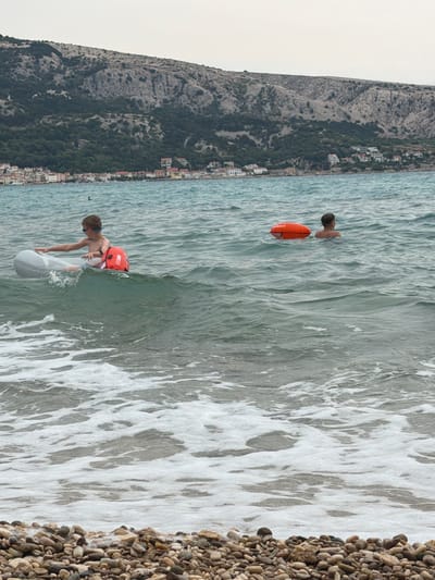The image shows a boy in the sea wearing an orange life vest and blue goggles. He is holding onto a white float or raft. In the background, another person is visible in the water with an orange float. The scene is set on a pebble beach with the sea to the right of the frame and a mountainous coastline with buildings in the background to the left. The water is choppy with waves breaking and creating foam. The sky appears overcast, suggesting a cloudy day. The location is identified as Općina Baška, Croatia. The boy's body language suggests he is actively engaged in swimming or playing in the water. The text "ASSISTO" is visible on the orange life vest.
