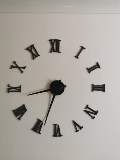 The image shows a close-up, eye-level, interior, medium shot of a black Roman numeral wall clock against a light-colored textured wall. The clock face is not visible, only the individual Roman numeral hour markers and the black clock hands. The Roman numerals are positioned around the clock face, with the hour hand pointing towards the VII (seven) and the minute hand pointing towards the XII (twelve). The clock appears to be a decorative, modern piece. The wall has some minor imperfections, such as a slight discoloration and some textured bumps. The lighting is even, suggesting artificial light or a well-lit room. There are no people or other objects visible in the frame. The location is not specified, but the scene depicts a domestic or commercial interior.
