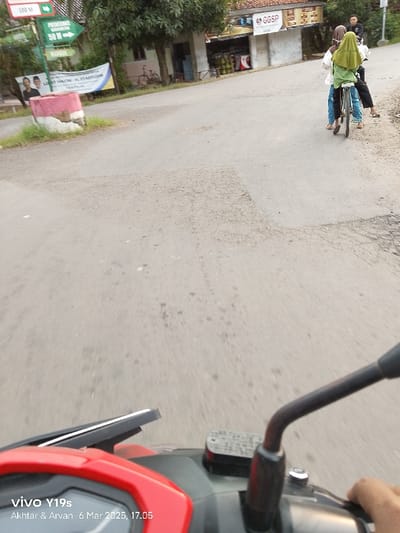 The image shows a view from a motorcycle traveling down a street. The road surface is gray asphalt with patches of missing pavement. There are two people on a bicycle in the distance, traveling in the same direction as the motorcycle. The motorcycle is red and black. The rider is wearing a black shirt and is holding the handlebars. The image was taken in the Kecamatan Gabuswetan area of Indonesia, at the coordinates -6.44 latitude and 108.078 longitude. The time is 6 March 2023, 17:05. On the left side of the road, there is a red-and-white pillar with a sign on it. The sign reads "Poskamling 50M" and "MAKMUR." The road is lined with green trees and buildings on both sides. The image was taken by a Vivo Y19s. A sign on the building on the right reads "GGSP."