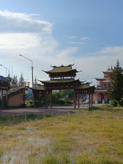This is a full, eye-level, outdoor, vertical, photographic shot of a Buddhist monastery entrance in Verkhnyaya Ivolga, Russia. In the foreground, a grassy field with patches of standing water occupies the lower half of the frame. The middle ground features a large, wooden, traditional East Asian-style gateway with multiple tiered roofs. To the left of the gateway, a wooden elevated walkway with stairs leads up to a building. Behind the gateway and walkway, a complex of buildings with pagoda-like roofs, painted in red and white, rises up. A large evergreen tree stands to the right of the buildings. The sky above is a clear blue with scattered white clouds. The lighting suggests it is daytime, possibly late afternoon. There are no people visible in the frame.