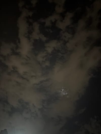 The image is a nighttime photograph taken from a low angle, looking up at a cloudy sky. The clouds are illuminated by an unseen light source, likely the moon or artificial lights, creating a contrast between dark shadows and lighter, hazy areas. There is a faint glimpse of what appears to be a building or structure at the very bottom left of the frame, silhouetted against the lighter sky. The overall atmosphere is dark and moody, with the clouds dominating the composition. There are no people or clear actions depicted in the scene. The specific location of Grapevine, United States of America, cannot be determined from the visual information provided in the image.