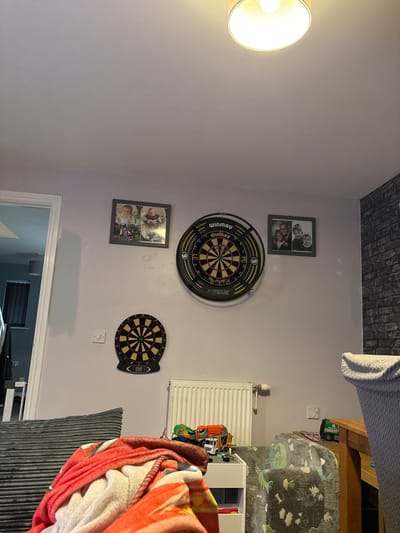 This image captures an interior domestic scene within the Forest of Dean, United Kingdom. The room features pale purple walls and a white ceiling illuminated by a central, drum-shaped ceiling light fixture. On the main wall, a large black Winmau Blade 6 Xtreme bristle dartboard with darts embedded is centrally mounted. Above and to its left, a framed photograph displays four smaller images, primarily featuring infants. To the right of the large dartboard, another framed photograph shows two adults and two children. A smaller, black electronic dartboard is positioned below the large dartboard and to its left, with a white light switch located adjacent to it.

A white panel radiator is affixed to the wall beneath the dartboards. The far right wall features dark grey brick-effect wallpaper, with a white electrical outlet or switch positioned low on this section. In the lower left foreground, a blanket primarily red, white, and blue in pattern is draped over an obscured object. To the right of the blanket, a low white shelf unit contains several toys, including a yellow digger, an orange dump truck, and a white truck. A grey armchair or sofa, patterned with green dinosaurs, is partially visible to the right of the shelf unit. Further to the right, a segment of a wooden table is visible, upon which a small green refuse truck toy rests. A white-trimmed doorway on the far left opens into an adjoining room, revealing darker walls and a vertical blind. No individuals are present.