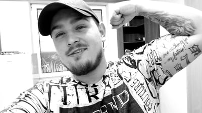 This black and white image captures a male individual indoors in Senec, Slovakia. The subject, framed from the chest up, faces the camera with a slight smile, wearing a dark baseball cap and a light-colored t-shirt featuring dark, abstract graphic patterns and text. He has a short beard and goatee. His left arm is raised and flexed, with the fist clenched, showcasing multiple tattoos. A full sleeve or large tattoo covers his upper left arm, while the inner bicep area displays text reading "is one authority for me - my father!" followed by stylized numerical tattoos, including "7" and "9". In the background to the left, a white "Amica" branded refrigerator or appliance is partially visible. Centrally, a window or glass panel is present, featuring a decorative etched or frosted botanical pattern, specifically depicting plant stalks, at its lower portion. To the right, dark shelving units are visible behind the subject's head and shoulder.