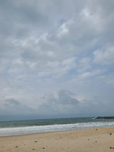 The image depicts a coastal scene on a cloudy day in Ibeju, Nigeria. The foreground is a light brown sandy beach, appearing damp near the waterline, with scattered dark debris and some small specks of green and white litter. The midground features the Atlantic Ocean with dark green to blue water, characterized by moderate waves breaking into white foam as they reach the shore. The waves exhibit a consistent pattern from left to right across the frame. On the far right, a dark grey rock jetty or breakwater extends from the land into the ocean, creating a barrier against the waves. The background consists of an overcast sky dominated by a large expanse of grey and white clouds, with small patches of lighter blue sky visible through breaks in the cloud cover, particularly towards the upper right. No individuals, vehicles, or specific activities are visible within the frame.