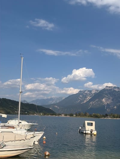 This is a full, vertical shot of a serene lake scene under a bright blue sky with scattered white clouds. The foreground is dominated by several docked white sailboats, their masts and rigging visible against the sky. One sailboat in the immediate foreground is partially covered with a light-colored tarp. Further out on the water, a small white motorboat with a covered cabin is anchored. The middle ground features the calm, blue water of the lake, with a few buoys floating near the docked boats. The background showcases a lush, green hillside transitioning into rugged, majestic mountains with visible striations and peaks. The shoreline is dotted with trees and buildings, suggesting a populated area. The overall impression is one of tranquility and natural beauty. The scene is likely Calceranica al Lago, Italy, a comune in the province of Trento in the Italian region of Trentino-Alto Adige/Südtirol, known for its picturesque lake.