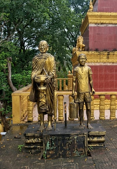A golden statue of a Buddhist monk stands to the left, holding a bowl in his hands and an umbrella leaning against his side. To his right is a golden statue of a young boy holding a basket. They are positioned on a dark base in front of a gold and red building with decorative elements. The statues are located outside, with lush green trees and foliage in the background. This scene is likely at a temple or religious site within Sihanoukville, Cambodia.
