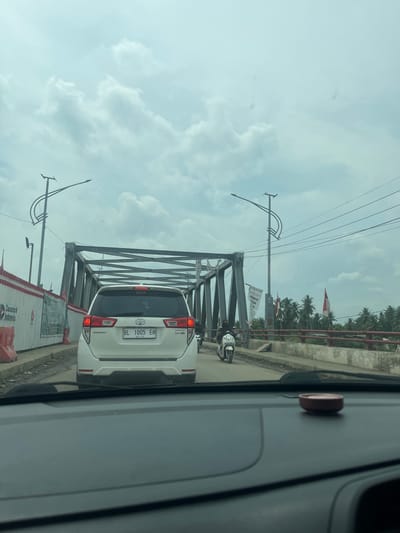 The image captures a traffic scene on a bridge, likely in Lhoknga, Indonesia. A white Toyota Innova vehicle with license plate "BL 1005 EO" is in the foreground, viewed from behind. To the left, a construction barrier with banners, including one with "Danantara Indonesia" and "Indonesia" is visible. A metallic bridge structure spans across the frame. A scooter with a rider is in motion on the bridge, heading towards the viewer. Palm trees line the background. The sky is overcast with clouds. The perspective suggests the photo was taken from inside another vehicle.